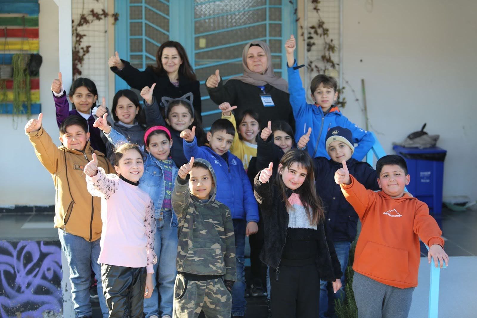 Group of students and teachers standing in front of a building while smiling and holding up their thumbs.